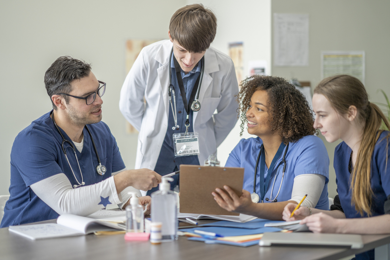 A multi ethnic group of nursing students are attending a meeting together with their male professor who is a doctor wearing a lab coat. They are seated at a table in a hospital and their professor is standing in between them while listening to their discussion.